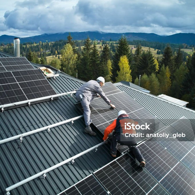 Zwei Monteure verlegen Solarmodule auf einem Blechdach, dahinter Wald